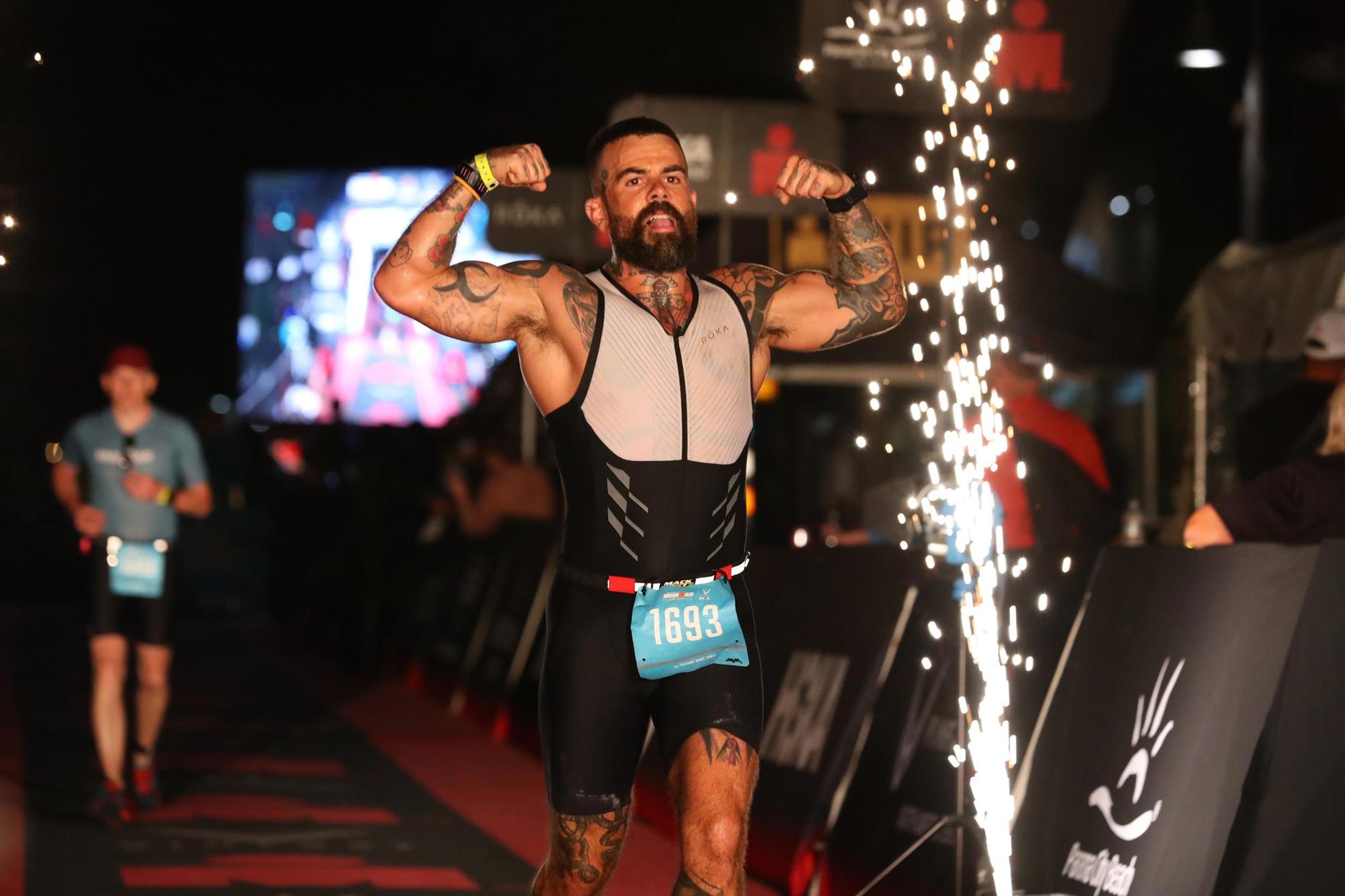 Scott Britton crossing the Ironman Florida finish line, arms raised, sparks behind